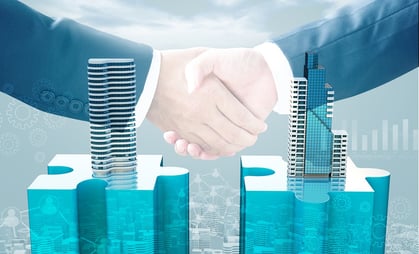 A photo of two hands shaking in agreement above two buildings coming out of puzzle pieces, indicating a successful merger.