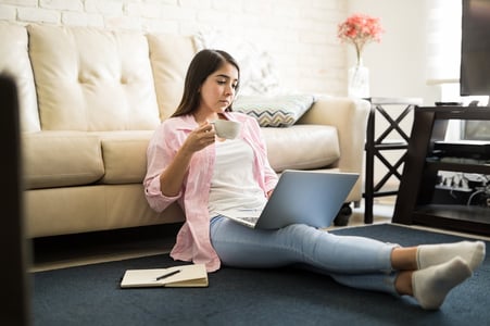 A photo of a woman sitting on the floor in her living room with her computer and a cup of coffee.