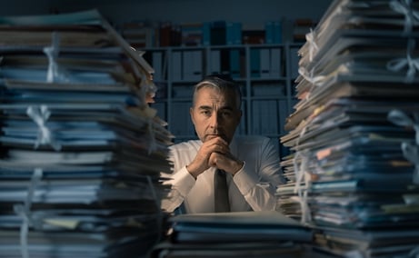 A photo of a man sitting in a darkened office staring at huge stacks of paperwork and file folders.