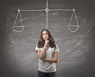 a photo of a woman standing in front of a chalkboard with a tipped scale on it with a thoughtful expression.