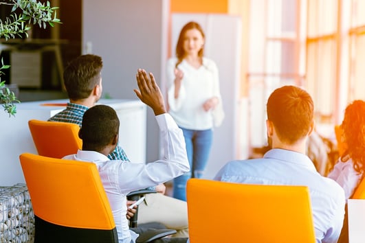 A photo of a man raising his hand to ask a presenter a question in an office.