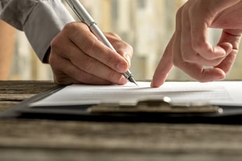 A close up photo of a person pointing to a line on a piece of paper and another person signing where indicated.