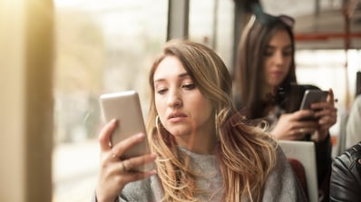 A photo of a young woman leaning against a window, looking at her smartphone.