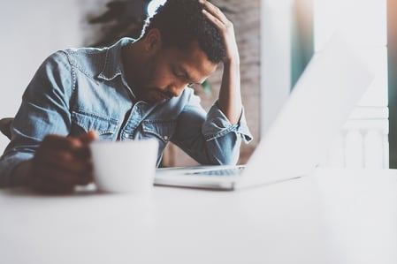 A photo of an exhausted man clutching a coffee cup in one hand while resting his head in the other hand.