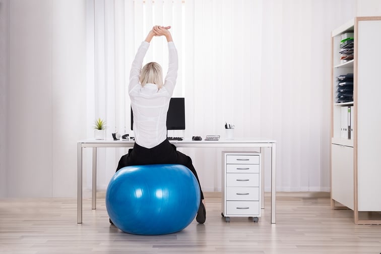 A photo of a woman sitting on a balance ball and stretching at her desk.